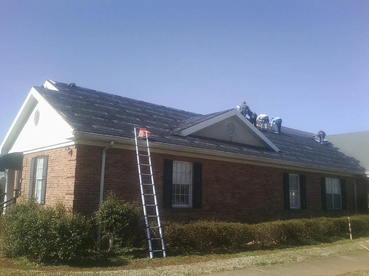 Crew tearing shingles off a brick ranch roof with a ladder set — Emergency Roof Repair work by our crew in Bonham